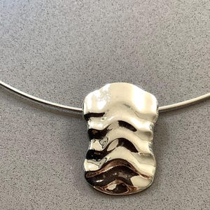 May include: A silver necklace with a pendant of an organic, textured form. The pendant's wavy surface reflects light, creating highlights and shadows. The necklace is presented on a grey surface.