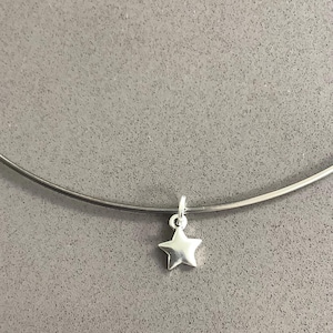 Puede incluir: Un collar gargantilla plateado con un pequeño colgante de estrella de cinco puntas. La estrella está centrada en el collar, que se encuentra sobre un fondo gris. El collar es una joya sencilla y elegante.