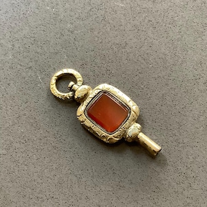 May include: A vintage gold-toned watch key with a rectangular, reddish-brown stone set in the center. The key has ornate detailing and a circular loop at the top.