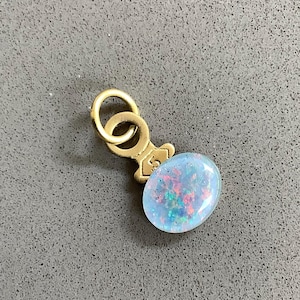 May include: A gold-toned metal pendant featuring a circular jump ring attached to a small, shield-shaped connector stamped with the number 5. The pendant is set with a smooth, oval-shaped cabochon displaying a light blue base with iridescent flecks.