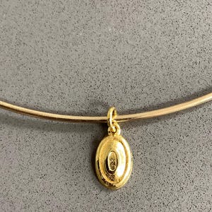 May include: A gold-toned necklace featuring a thin, rigid circular band and a single oval-shaped pendant. The pendant has a raised, centred oval detail and is attached to the band by a small, simple gold-toned jump ring loop.