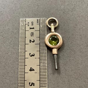 May include: A gold-toned watch key with a round, olive-green gemstone set in the center. The key is approximately 1 inch long, with a small loop at the top and a slender, dark-colored stem.