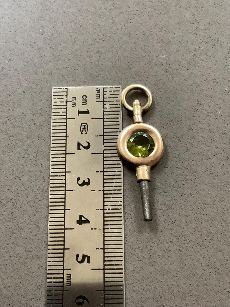 May include: A gold-toned watch key with a round, olive-green gemstone set in the center. The key is approximately 1 inch long, with a small loop at the top and a slender, dark-colored stem.