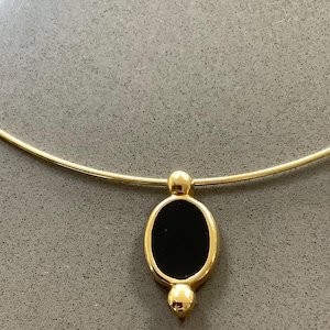 May include: A gold-toned necklace with a black oval pendant. The pendant is framed in gold and has a small gold sphere above and below the oval. The necklace is on a gray background.