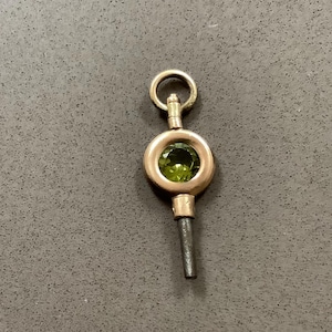 May include: A vintage gold-toned watch key with a circular design. The key features a green gemstone set in the center and a small loop at the top. The stem is a dark gray color.
