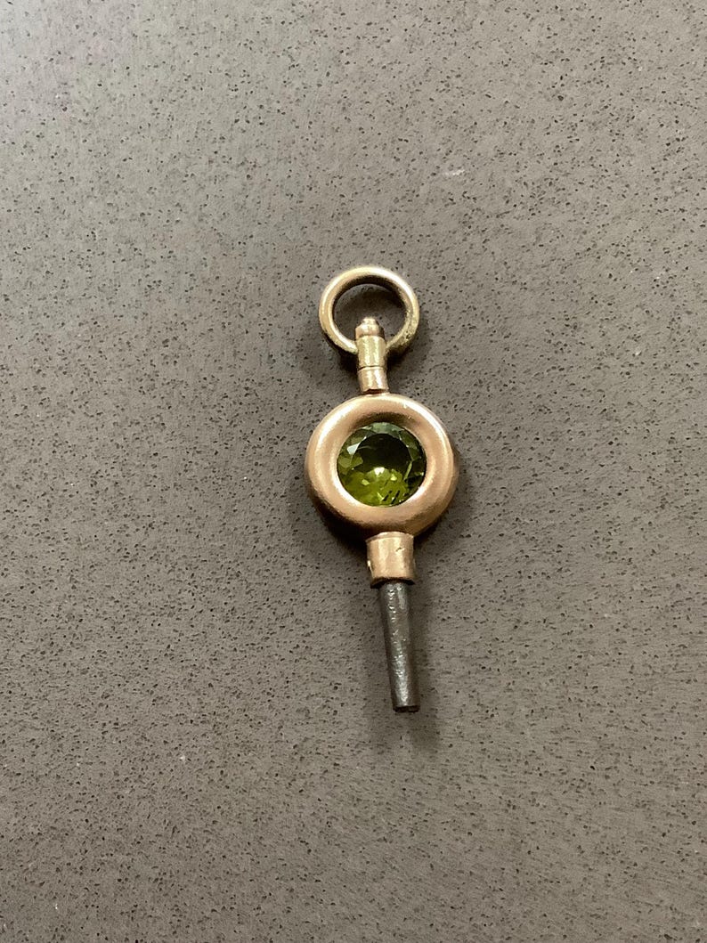 May include: A vintage gold-toned watch key with a circular design. The key features a green gemstone set in the center and a small loop at the top. The stem is a dark gray color.
