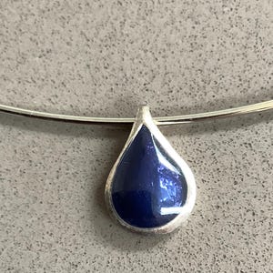 May include: A teardrop-shaped pendant featuring a deep, glossy blue enamel inlay set within a smooth silver-toned metal frame. The pendant is suspended from a thin, rigid silver-toned metal collar necklace that curves gently across the top.