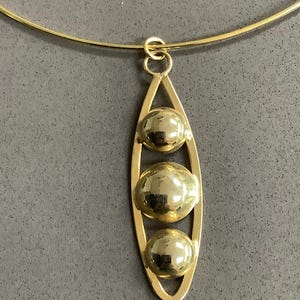 May include: A gold-coloured necklace with a pendant. The pendant has three textured, spherical orbs within an elongated, vertical frame. The necklace features a thin, gold-toned choker-style chain.