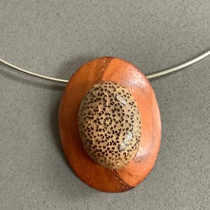 May include: A handcrafted necklace with a wooden pendant. The pendant has an oval shape, with a speckled stone set on top of an orange-brown wooden base. The necklace chain is a thin, silver-coloured wire. The pendant is approximately 5 cm in length.