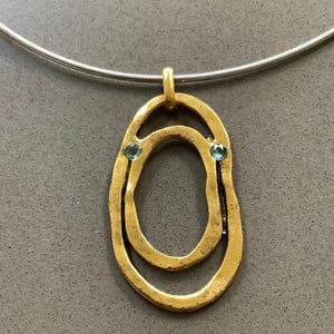 May include: A gold-coloured pendant necklace with a double oval design and a textured surface. The pendant has two small blue gemstones and hangs from a silver-coloured choker-style chain. The necklace is displayed against a grey background.
