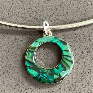 May include: A circular pendant featuring a vibrant, iridescent green and blue abalone shell inlay set within a metallic frame. The pendant hangs from a simple, smooth silver-toned metal collar necklace, creating a clean and reflective jewellery piece.