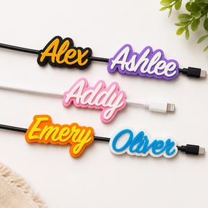 May include: Five custom cable identifiers featuring the names Alex, Ashlee, Addy, Emery, and Oliver in various colors including yellow, white, pink, orange, and blue, attached to black and white charging cables with USB-C and Lightning connectors.