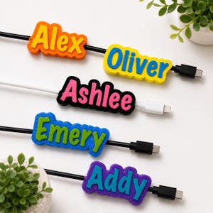 May include: Four custom cable identifiers featuring the names Alex, Oliver, Ashlee, Emery, and Addy in raised, colourful 3D lettering. Each tag is attached to a charging cable, showcasing combinations like orange and yellow, yellow and blue, black and pink, blue and green, and purple and teal.