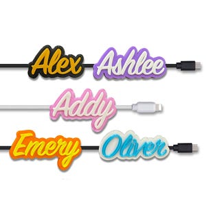 Puede incluir: Una colección de organizadores de cables personalizados, cada uno con un nombre en una fuente de burbujas colorida. Los nombres incluyen "Alex", "Ashlee", "Addy", "Emery" y "Oliver". Cada organizador está conectado a un cable de carga con un conector USB o Lightning.