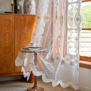 Puede incluir: Un panel de cortina blanco transparente con intrincados bordados en tono dorado en patrones ovalados y florales. La tela tiene un borde inferior festoneado con un delicado diseño de celosía. La cortina está drapeada sobre una mesa de madera.