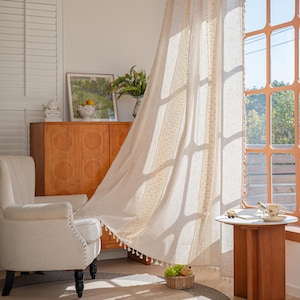 May include: Beige linen curtain with a decorative lace trim and tassels. The curtain is hanging in front of a window, with sunlight streaming through. A white armchair and wooden cabinet are in the background.