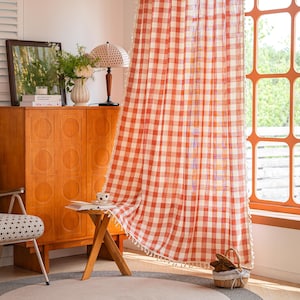 May include: A floor-length curtain featuring a red and white gingham check pattern. The fabric is semi-sheer and includes small white decorative tassels along the vertical edges. The curtain hangs from a rod and covers a portion of a window.