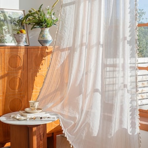 以下が含まれることがあります： 垂直ストライプとタッセルトリムのシアーホワイトカーテン。カーテンは窓の前に吊り下げられ、太陽光が差し込んでいます。小さな丸いテーブルにはティーカップと本が置かれています。木製のキャビネットと花瓶も表示されています。