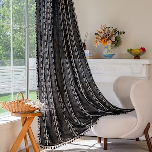 May include: Black sheer curtain with a geometric pattern of squares and vertical stripes. The curtain has a fringe of small black tassels along the bottom edge. The curtain is hanging in front of a window, with a view of a building and trees.