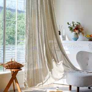 以下が含まれることがあります： 大きな窓の前に吊るされたベージュと白のストライプのカーテンパネル。カーテンは質感があり、タッセルでトリミングされています。窓の前には、籐のバスケットが付いた木製のサイドテーブルがあります。