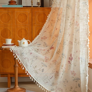 May include: A sheer, off-white curtain panel featuring a delicate botanical print of wildflowers in muted pink, blue, and green tones. The fabric has a visible woven texture and is finished with small, white decorative tassels along the edge.