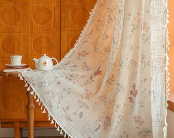 May include: A sheer, off-white curtain panel featuring a delicate botanical print of wildflowers in muted pink, blue, and green tones. The fabric has a visible woven texture and is finished with small, white decorative tassels along the edge.