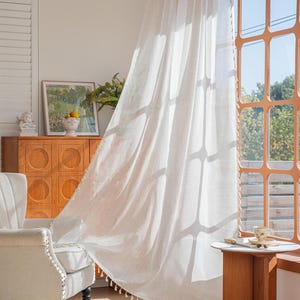 Pode incluir: Cortina branca transparente soprando em frente a uma janela com moldura de madeira. Uma pequena mesa de madeira suporta uma xícara e um pires. Uma cesta de vime com frutas está no chão. Uma poltrona clara está em primeiro plano.