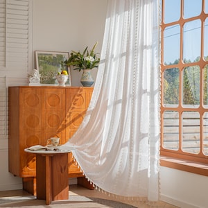 以下が含まれることがあります： 窓の前に、繊細な模様とタッセルのトリムが付いた、透け感のある白いカーテンが吊るされています。 カーテンは部分的に開いており、外の景色が見えます。 木製のキャビネットと小さなテーブルが手前にあります。