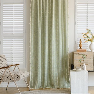 May include: A sage green curtain with vertical white stripes and a fringed edge hangs in front of a window. The curtain is made of a textured fabric and is hanging from a black rod. The room has white shutters and a patterned chair.
