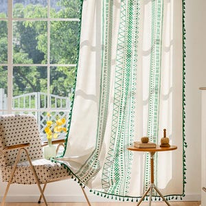 Op de afbeelding: Witte linnen gordijnen met groene geometrische borduursels en kwastjes. De gordijnen hangen voor een raam, met een stoel met patroon en een kleine tafel zichtbaar. Het ontwerp heeft verschillende patronen.