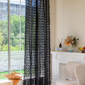 May include: Black sheer curtain with a geometric pattern and tassel trim. The curtain hangs in front of a window, with a view of trees and a white frame. The curtain is part of a home decor collection.