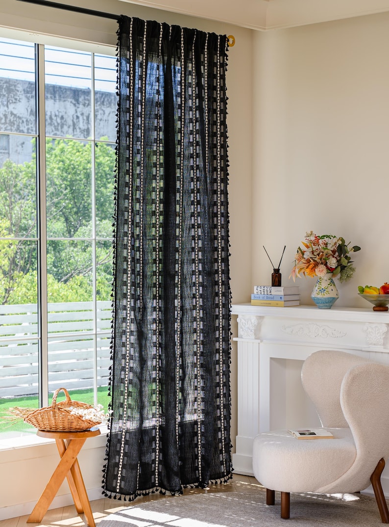 May include: Black sheer curtain with a geometric pattern and tassel trim. The curtain hangs in front of a window, with a view of trees and a white frame. The curtain is part of a home decor collection.