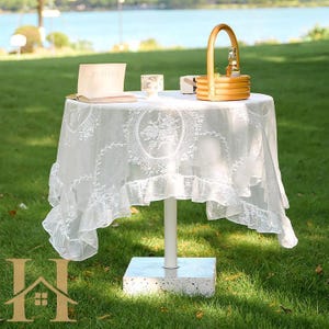 May include: A white, round table with a delicate lace tablecloth featuring floral patterns and a ruffled edge. A small picnic basket, a book, and a cup sit on the table. The table is supported by a white pole and a square concrete base, set on a grassy lawn.