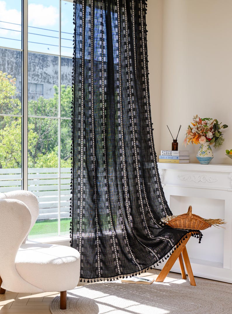 May include: Black sheer curtain with a geometric pattern of white squares and vertical stripes. The curtain has a fringe of small black tassels along the edges. The curtain hangs in front of a large window, with a white chair and a small wooden stool in the foreground.