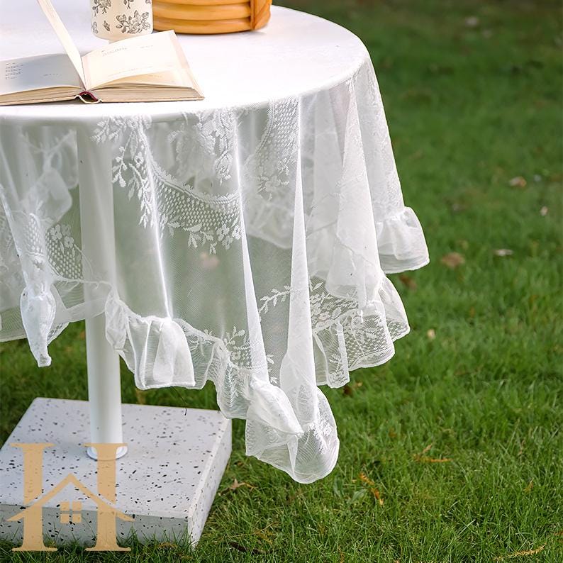 May include: A white lace tablecloth draped over a round table. The tablecloth features a floral lace pattern and a ruffled edge. An open book and a decorative item sit on the table. The table is on a grassy surface.
