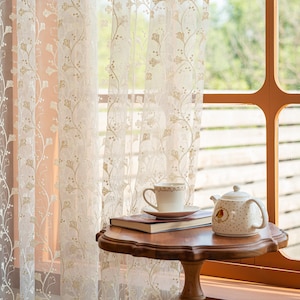 May include: Sheer white curtains featuring a delicate gold embroidered ginkgo leaf pattern hang in front of a window. A wooden pedestal table holds a white ceramic teacup on a saucer, a closed book, and a white teapot with a small bird illustration.