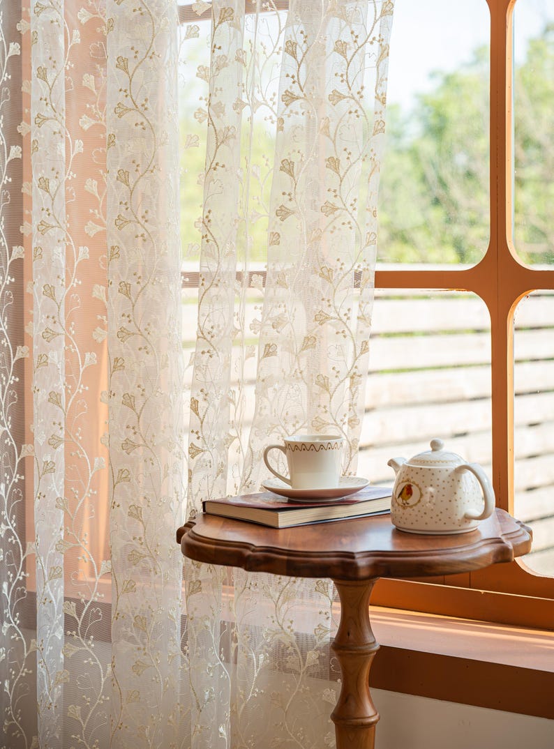 May include: Sheer white curtains featuring a delicate gold embroidered ginkgo leaf pattern hang in front of a window. A wooden pedestal table holds a white ceramic teacup on a saucer, a closed book, and a white teapot with a small bird illustration.