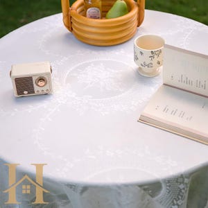 May include: A white round tablecloth with a floral design, set with a small radio, a cup with floral patterns, a book, and a basket. The basket contains a bottle and a pear. The scene is set outdoors.