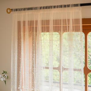 May include: A sheer, off-white curtain panel featuring a delicate, repeating embroidered pattern of small, light-colored botanical sprigs and leaves. The lightweight fabric hangs from a dark rod with gold-toned end caps, creating soft folds.
