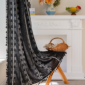 May include: Black curtain with white geometric patterns and tassels. The curtain is draped over a wooden stool. A basket of dried flowers, a vase of flowers, and a bowl of fruit are on a white mantelpiece.