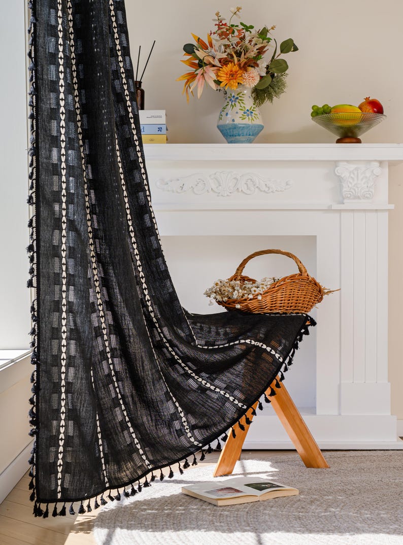May include: Black curtain with white geometric patterns and tassels. The curtain is draped over a wooden stool. A basket of dried flowers, a vase of flowers, and a bowl of fruit are on a white mantelpiece.