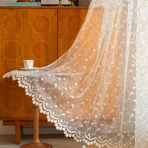May include: A sheer white curtain panel featuring an intricate embroidered pattern of delicate vines and ginkgo-like leaves. The fabric has a scalloped bottom edge finished with a detailed lace trim, creating a soft and translucent appearance.