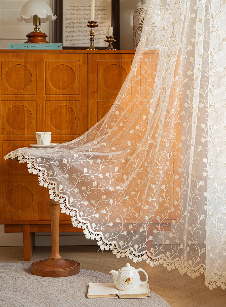 May include: A sheer white curtain panel featuring an intricate embroidered pattern of delicate vines and ginkgo-like leaves. The fabric has a scalloped bottom edge finished with a detailed lace trim, creating a soft and translucent appearance.