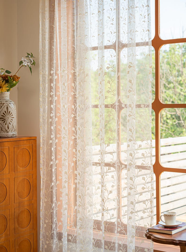 May include: A sheer white curtain panel featuring a delicate, repeating vertical pattern of embroidered light brown vines and small leaves. The lightweight fabric allows soft natural light to filter through, highlighting the intricate botanical design.