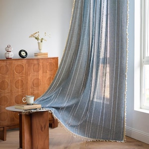 以下が含まれることがあります： 垂直の白いストライプとクリーム色のフリンジが付いた、薄い青色のカーテン。カーテンは窓の前に吊り下げられており、手前に木製のキャビネットと小さなテーブルがあります。テーブルの上にはカップと本があります。