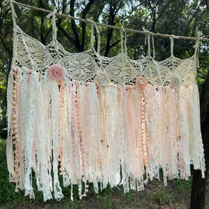 Könnte beinhalten: Ein cremefarbener, handgefertigter Spitzenvorhang mit einem gewellten oberen Rand. Der Vorhang hat hängende Bänder in Creme und Rosa, die mit kleinen rosa Pompons und Stoffrosen verziert sind. Der Vorhang ist an einem Naturseil aufgehängt.