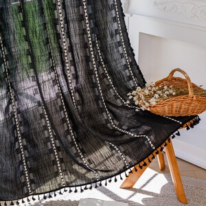 May include: Black sheer curtain with a geometric pattern of white squares and vertical lines. The curtain has a tassel trim along the bottom edge. A woven basket with dried flowers sits on a wooden stool. An open book rests on the floor.