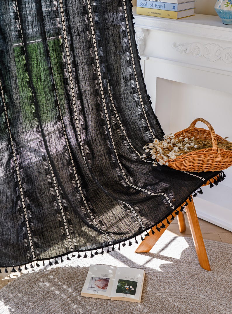 May include: Black sheer curtain with a geometric pattern of white squares and vertical lines. The curtain has a tassel trim along the bottom edge. A woven basket with dried flowers sits on a wooden stool. An open book rests on the floor.