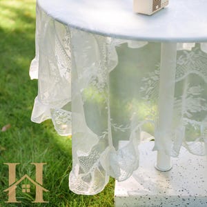 May include: A white, round table covered with a delicate, lacy tablecloth. The tablecloth features floral patterns and a ruffled edge, creating an elegant and romantic aesthetic. The table is set outdoors on a grassy surface.