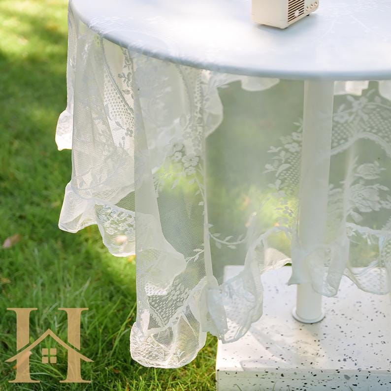 May include: A white, round table covered with a delicate, lacy tablecloth. The tablecloth features floral patterns and a ruffled edge, creating an elegant and romantic aesthetic. The table is set outdoors on a grassy surface.
